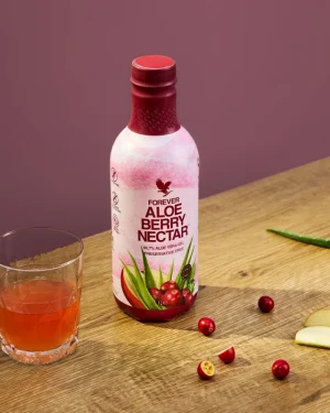 Forever Aloe Berry Nectar bottle on wooden table beside a glass of pink aloe drink, cranberries and apple slices.
